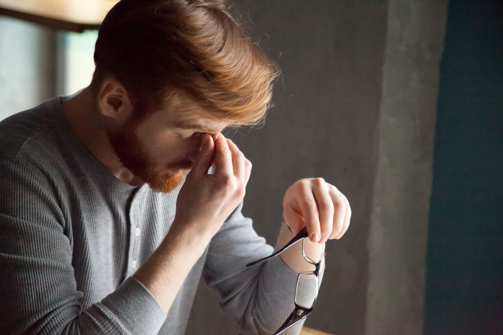 A man at a desk with his eyes closed, holding his spectacles in one hand and pinching the bridge of his nose with the other